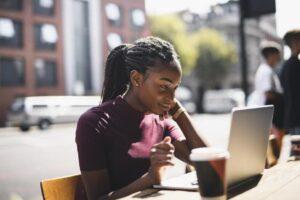 Eine schwarze CBD-Unternehmerin in einem sonnigen Café, die lächelnd auf ihren Laptop schaut, während sie sich Facebook-Marketing-Tipps ansieht.