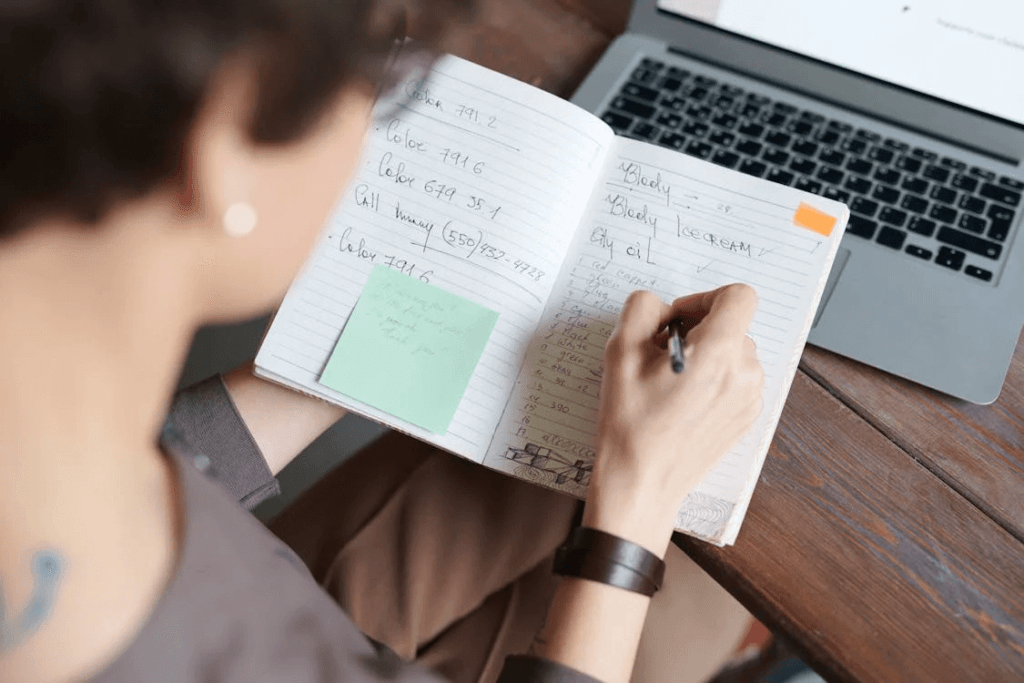 A person writing notes in a notebook with a sticky note attached, next to a laptop.